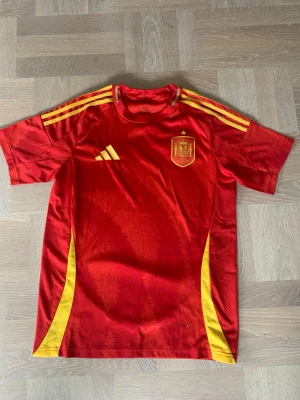 Spaniens matchtröja Adidas röd/gul - Säljer en officiell röd matchtröja från Spaniens landslag, tillverkad av Adidas. Tröjan har gula detaljer på sidorna och axlarna, samt landslagets emblem på bröstet. Tillverkad i lätt och ventilerande material, perfekt för fotboll eller träning. Spaniens EM 2024 tröja. Fint skick använd fåtal gånger, storlek S.