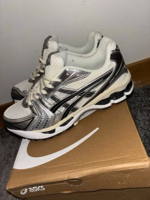 asics gel kayano 14 - Snygga sneakers från ASICS i vitt och silver med svarta detaljer. Ovandelen är i mesh och syntetmaterial för bra andningsförmåga och komfort. Klassisk snörning och chunky sula ger en sportig vibe. Perfekta för dig som gillar retrostil och streetwear.