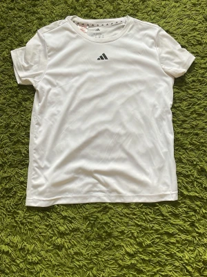 Vit Adidas t-shirt i polyester - Säljer en klassisk vit t-shirt från Adidas med diskret logga på bröstet. Tillverkad i mjuk polyester som andas, perfekt för träning eller chill. Enkel och stilren design med rund hals och korta ärmar.