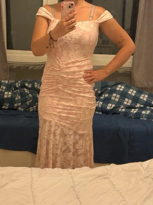 Ljusrosa spetsklänning med offshoulder -  långklänning i ljusrosa spets med offshoulder och figurnära passform. Klänningen har vackra blommiga spetsdetaljer, lätt rynkad midja och är tillverkad i ett mjukt syntetmaterial. Perfekt för sommarens speciella tillfällen.