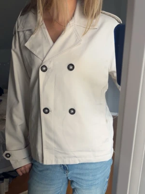 Beige skepparkavaj med svarta knappar - Snygg beige trenchcoat med dubbelknäppning och svarta knappar. Rak passform. Passar även mig som brukar ha S
