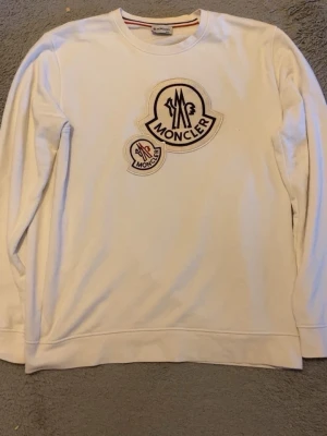 Vit sweatshirt från Moncler XXL - Vit sweatshirt från Moncler med stora broderade loggor på bröstet. Tröjan har rund halsringning, långa ärmar och ribbade muddar. Klassisk och stilren design med exklusiv känsla, perfekt för dig som gillar streetwear och premiumvarumärken.