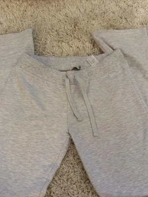 Ljusgrå mjukisbyxor från Nelly - Säljer ett par ljusgrå mjukisbyxor från Nelly med dragsko i midjan och ribbade muddar. Byxorna är tillverkade i mjukt bomullsmaterial och har en relaxed fit som är perfekt för chill dagar. Klassisk design utan tryck eller detaljer.