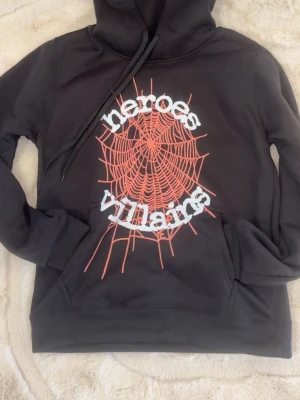 Svart hoodie Heroes Villains tryck - Svart hoodie med stor röd spindelnätsgrafik och texten 'heroes villains' i vitt på framsidan. Klassisk känguruficka och huva med dragsko. Tillverkad i mjukt bomullsmaterial, perfekt för en avslappnad streetwear-look.