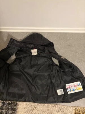 Svart dunväst från Moncler med luva - Snygg svart dunväst från Moncler med avtagbar luva och två dragkedjefickor framtill. Västen har ett quiltat mönster och klassisk Moncler-logga på bröstet. Perfekt för lager-på-lager och streetstyle.