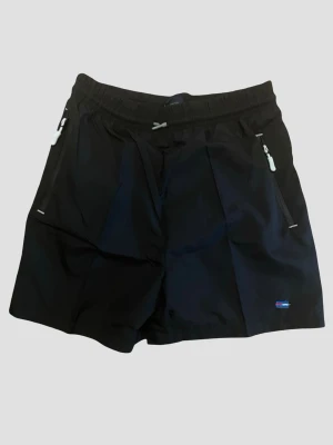 Badshorts från Tommy Hilfiger  - Snygga marinblå badshorts från Tommy Hilfigsr  med elastisk midja och dragsko. Shortsen har två dragkedjeförsedda fickor fram och en reflexdetalj bak. Perfekta för sommarens alla äventyr och har en sportig look.