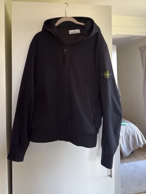 Stone Island jacka  - Svart vindjacka från Stone Island med huva och dragkedja framtill. Jackan har det klassiska Stone Island-märket på vänster ärm och är tillverkad i Light Soft Shell-R material som är lätt och vindtätt. Två sidofickor med dragkedja och ribbade muddar vid ärmslut och nederkant. Öppen för prisförslag