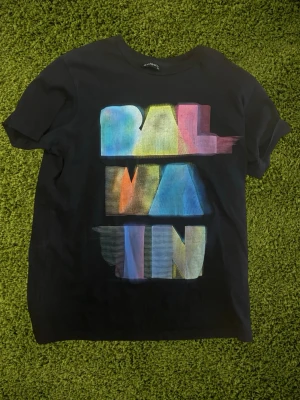 Svart Balmain t-shirt med färgglatt tryck - Cool svart t-shirt från Balmain i storlek XL (liten i storleken: motsvarar L i verkligheten) med ett stort färgglatt grafiskt tryck på framsidan där det står BALMAIN. T-shirten har rund hals och korta ärmar. Perfekt för dig som vill sticka ut med en unik design.