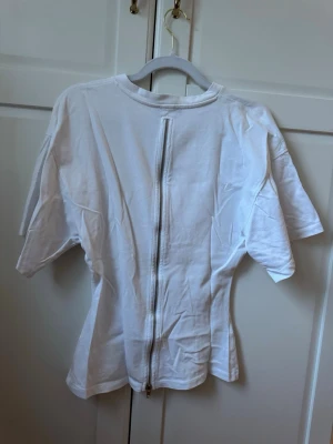 Vit t-shirt med dragkedja bak - Vit t-shirt i bomull med rund hals och korta ärmar. Unik detalj med en lång dragkedja längs ryggen som ger en cool twist till en klassisk look. Passformen är normal och tyget känns mjukt och bekvämt.