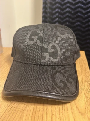 Svart keps från Gucci med GG-mönster - Snygg svart keps från Gucci med det klassiska GG-mönstret i grått över hela kepsen. Kepsen har böjd skärm och justerbart spänne bak. Tillverkad i slitstarkt tyg med exklusiv känsla, perfekt för dig som vill ha en stilren accessoar.
