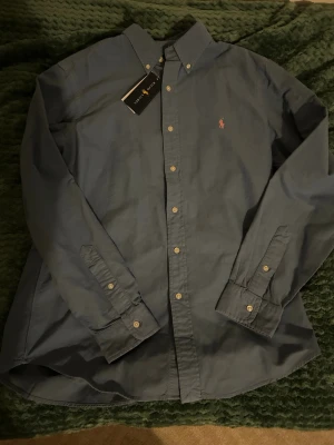 Blå skjorta från Ralph Lauren - Klassisk blå skjorta från Ralph Lauren med button-down krage och den ikoniska rosa broderade loggan på bröstet. Skjortan har långa ärmar och knappar framtill. Tillverkad i mjuk bomull för en bekväm känsla.