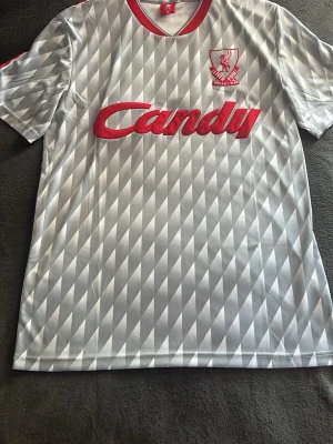 Liverpool Candy retro bortatröja grå/röd - Retroinspirerad Liverpool FC bortatröja i grått med geometriskt mönster och röda detaljer. Tryckt Candy-logga på bröstet och klubbmärke i rött. Kortärmad och tillverkad i lätt, ventilerande polyester – perfekt för fotboll eller träning.