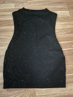 Svart glittrig kortklänning - Snygg svart kortklänning med glittrigt mönster som ger en stjärnhimmel-vibe. Klänningen har en figurnära passform och är ärmlös, perfekt för dig som vill sticka ut. Materialet känns mjukt och stretchigt, troligen polyester eller syntet.