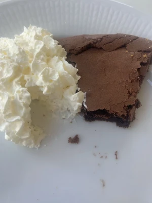 Kladdkaka med grädde - En bit kladdkaka med krispig yta och seg insida, serverad med fluffig vispgrädde på en vit tallrik. Perfekt för dig som älskar chokladiga bakverk och vill ha något riktigt gott till fikat.
