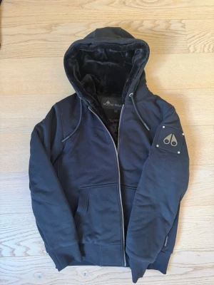 Svart hoodie från Moose Knuckles - Svart hoodie från Moose Knuckles med dragkedja, huva och mjukt foder. Snygg logotyp i metall på ärmen och praktiska fickor framtill. Perfekt för dig som gillar streetwear och vill ha något både stilrent och bekvämt.