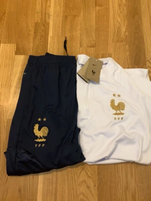 Nike FFF träningsset vitt och blått - Säljer ett träningsset från Nike med FFF-logga. Setet består av en vit långärmad tröja med gulddetaljer, halv dragkedja och mönstrade partier på ärmarna samt ett par mörkblå byxor med guldfärgad tupp och FFF-tryck. Perfekt för fotbollsträning eller chill.