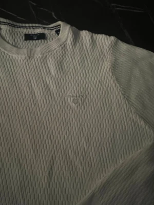 Vit stickad tröja från Gant - Klassisk vit stickad tröja från Gant med rund halsringning och diskret logga broderad på bröstet. Tröjan har lång ärm och ribbade muddar vid ärmslut och nederkant. Tillverkad i mjukt material som ger en clean och stilren look.