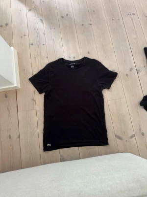 Svart t-shirt från Lacoste - Säljer en svart t-shirt från Lacoste med klassisk rund hals och korta ärmar. T-shirten har den ikoniska krokodilloggan broderad längst ner på framsidan. Perfekt för dig som gillar stilrena plagg med en touch av premium.