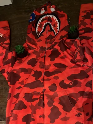 Bape hoodie - Säljer en ikonisk röd camo hoodie från BAPE med hajmotiv på huvan och broderade bokstäver i blått, orange och rött. Hoodien har dragkedja framtill och två fickor. Perfekt statement-plagg för streetwear-stilen. Vet ej om den e äkta