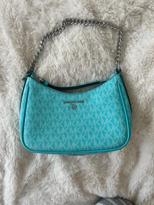 Turkos Michael Kors handväska - Snygg turkos handväska från Michael Kors med silverfärgad kedjerem och diskret MK-mönster över hela väskan. Väskan har en mjukt rundad form och detaljer i konstläder. Perfekt accessoar för att lyfta din outfit.