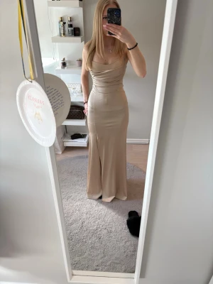 Champagnefärgad långklänning med snörning - Elegant långklänning i champagnefärg med smickrande drapering och snörning i ryggen. Klänningen har smala axelband, öppen rygg och en figurnära passform. Perfekt för speciella tillfällen när du vill sticka ut.  Köpt för 699kr