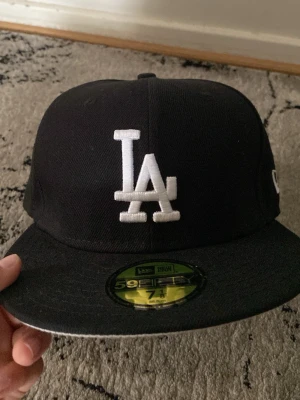 Svart LA New Era 59Fifty keps - Säljer en svart New Era 59Fifty keps med LA-logga broderad i vitt framtill och New Era-logga på sidan. Klassisk flat brim och stilren design, perfekt för dig som gillar streetwear. Materialet är bomull och kepsen har en strukturerad passform.