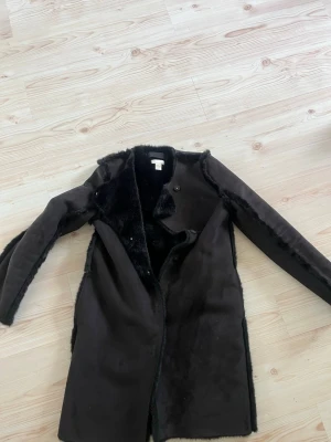 Svart fuskpälsfodrad kappa H&M - Snygg svart kappa från H&M med mjukt fuskpälsfoder på insidan. Kappan har en rak passform, långa ärmar och stängs med knappar framtill. Perfekt för kalla dagar när du vill vara både varm och stilren.