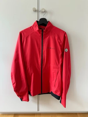 Röd vindjacka från Sail Racing - Snygg röd vindjacka från Sail Racing med svart dragkedja och detaljer. Jackan har hög krage, logga på bröstet och på ärmen. Tillverkad i lätt och vindtätt material, perfekt för blåsiga dagar. Sportig passform och elastiska muddar.