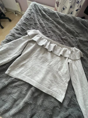 Ljusgrå offshoulder topp med volang - Superfin ljusgrå offshoulder topp från H&M Divided med bred volang runt axlarna. Mjuk och skön i bomullsmix, perfekt till jeans eller kjol. Långärmad och med en snygg croppad känsla. Enkelt att styla för en trendig look.