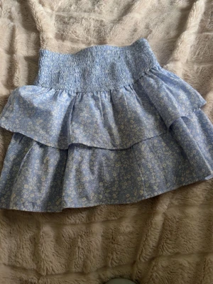 Blommig kort  volangkjol i ljusblått - Supersöt kort kjol i ljusblått med små vita blommor och bred smock i midjan. Kjolen har dubbla volanger som ger en luftig och lekfull känsla. Perfekt för varma dagar och enkel att matcha med en enkel topp.