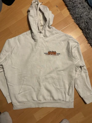 Vit hoodie från Chronike med tryck - Vit hoodie från Chronike med trycket 'Your Time Is NOW' i orange och svart på bröstet. Klassisk känguruficka framtill, huva med snörning och ribbade muddar. Perfekt för en chill och avslappnad stil.