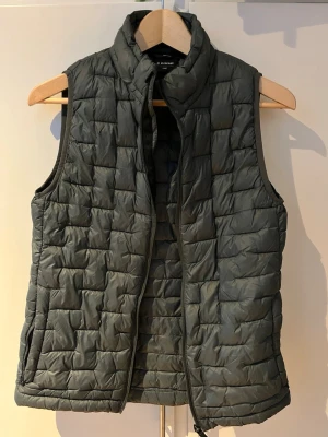 Olivgrön väst - Olivgrön väst från Everest med quiltat mönster. Västen har dragkedja framtill och två sidofickor. Perfekt att slänga över en hoodie eller tröja för extra värme. Tillverkad i lätt och smidigt material med polyesterfyllning.