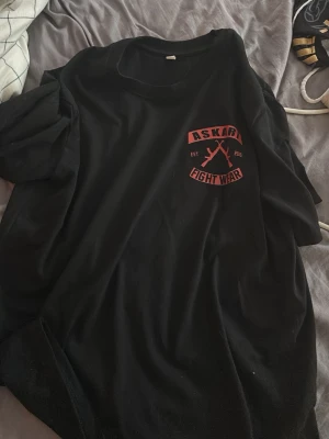 Svart Askari Fight Wear t-shirt - Svart t-shirt från Askari Fight Wear med röd logga och text både fram och bak. Trycket visar två korsade gevär och texten 'ASKARI FIGHT WEAR'. Klassisk rund hals och korta ärmar. Perfekt för dig som gillar streetwear och sportig stil.
