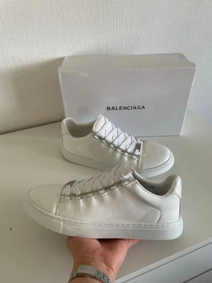 Vita Balenciaga arenas i läder - Stilrena vita sneakers från Balenciaga i slätt läder med vita skosnören och diskret design. Klassisk låg modell med tjock sula och metalliska detaljer vid snörningen. Perfekta för dig som gillar minimalistisk och exklusiv stil.