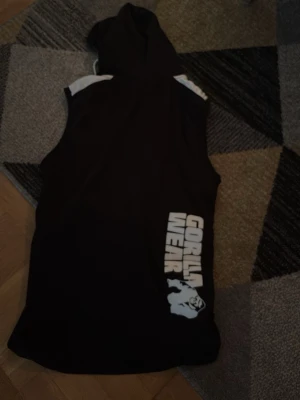 Svart ärmlös hoodie Gorilla Wear - Säljer en svart ärmlös hoodie från Gorilla Wear med vita detaljer och tryck på både bröstet och ryggen. Perfekt för gymmet eller träning, med huva och fickor framtill. Materialet känns mjukt och stretchigt, troligen polyester eller bomullsblandning.