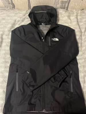 Svart regnjacka från The North Face - Snygg svart regnjacka från The North Face med huva och dragkedja framtill. Jackan har två sidofickor med dragkedja och meshfoder på insidan för extra ventilation. Perfekt för regniga dagar och enkel att matcha med allt. XXL för barn