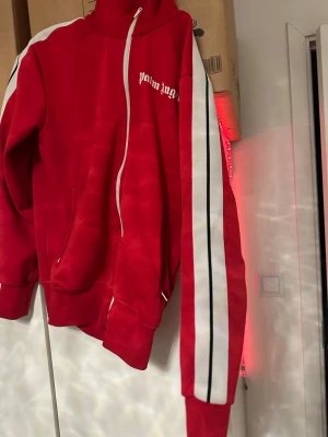 Röd Palm Angels hoodie med vita ränder - Säljer en röd hoodie från Palm Angels med vita och svarta ränder längs ärmarna. Dragkedja framtill, hög krage och fickor på sidorna. Palm Angels-logga i vitt på bröstet. Perfekt för en streetwear-look.