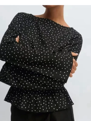 Svart prickig blus med volang - Trendig svart blus med vita små prickar, långärmad och med smockad midja samt volang nertill. Blusen har en rund halsringning och vida ärmar, vilket ger en snygg och avslappnad siluett. Perfekt för dig som gillar en lekfull men stilren look.