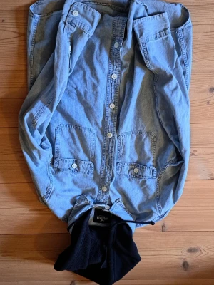 Ljusblå jeansjacka med huva - Snygg ljusblå jeansjacka från Hollister med svart huva i sweatshirtmaterial. Jackan har två stora bröstfickor med lock och knappar framtill. Klassisk rak passform och vita knappar ger en clean look.