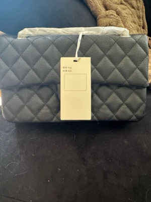 Svart quiltad handväska - Stilren svart handväska med quiltat mönster och rektangulär form. Väskan har en ficka på baksidan och ett lock framtill. Tillverkad i lambskin med struktur. Lång axelrems kedja. Perfekt för dig som gillar klassisk och trendig design.