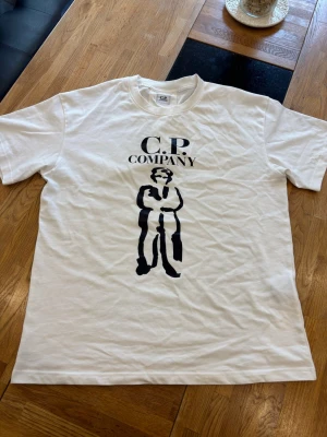Vit C.P. Company t-shirt - Säljer en vit t-shirt från C.P. Company med ett stort svart tryck av en person och loggan på bröstet. Klassisk passform och rund hals. Perfekt för dig som gillar streetwear och vill ha något stilrent men ändå med attityd.