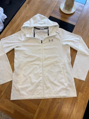 Under Armour Windbreaker  - Snygg vit vindjacka från Under Armour med huva och hel dragkedja framtill. Jackan har två sidofickor och ett diskret logotryck på bröstet. Tillverkad i lätt och tunt material, perfekt för blåsiga dagar. Sportig design med meshfoder för extra komfort.