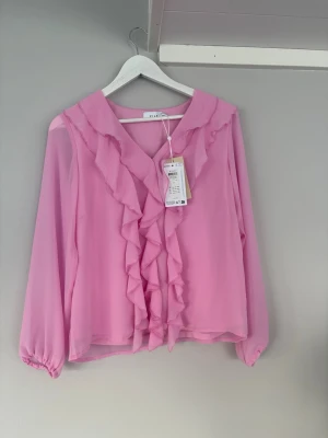 Rosa volangblus från Vila - Supersöt rosa blus från Vila med v-ringning och volangdetaljer längs halsringningen och framtill. Blusen har långa ärmar med resår vid ärmslut och är tillverkad i ett lätt, transparent material som ger en luftig känsla. Oandvänd!