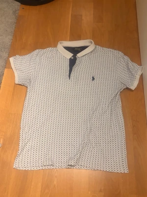Vit mönstrad t-shirt från Polo Ralph Lauren - Säljer en vit t-shirt från Polo Ralph Lauren med små blå mönster över hela tröjan. T-shirten har korta ärmar, blå knappslå . Använt för golf men funkar som vanligpike också sitter som S även om de är M
