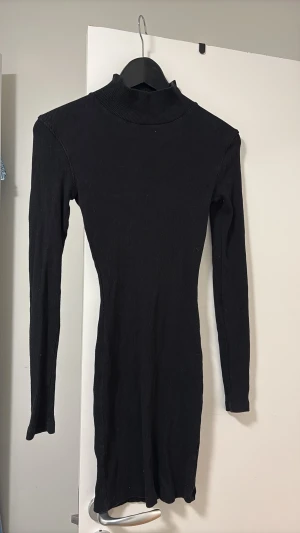 Svart ribbad poloklänning - Säljer en svart, ribbad kortklänning med lång ärm och polokrage. Klänningen har en tight passform och är perfekt för dig som gillar en enkel men stilren look. Materialet känns mjukt och stretchigt, vilket gör den bekväm att bära hela dagen.