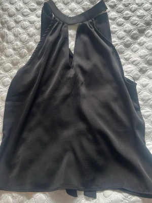 Svart halterneck linnetopp i satin - Snygg svart halterneck linnetopp i satin med knytband i nacken och en liten slits framtill. Toppen har en elegant, glansig finish och är ärmlös med lös passform. Perfekt för dig som vill ha en stilren och trendig topp.