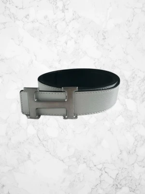 Vitt skärp med silverfärgat H-spänne - Snyggt vitt skärp med ett stort, silverfärgat H-format spänne. Skärpet har en stilren och modern look med slät yta och kontrastsömmar längs kanterna. Perfekt accessoar för att lyfta din outfit.