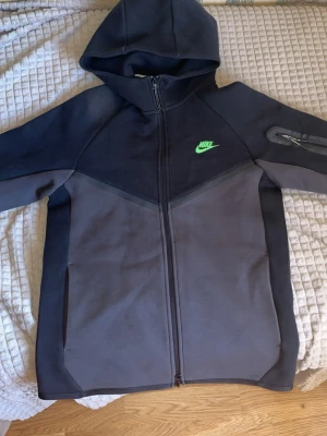 Nike Tech Fleece hoodie svart/grå - Nike Tech Fleece hoodie i svart och mörkgrått med hel dragkedja och huva. Har en grön Nike-logga på bröstet och en praktisk ficka med dragkedja på ärmen. Tillverkad i mjukt och bekvämt material som passar perfekt till streetwear-stilen.