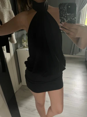Svart halterneck kortklänning - Säljer en svart kortklänning med halterneck och öppen rygg. Klänningen har en lös passform upptill och sitter tightare över höfterna. Perfekt för dig som vill ha en stilren och trendig look.