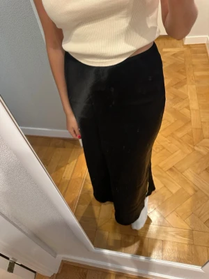 Svart långkjol i satin  - Stilren svart långkjol i lätt glansig satin. Kjolen har en rak passform och en liten slits på sidan för extra rörelsefrihet. Från Arket och aldrig använd!
